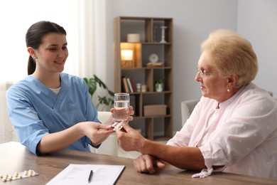 Photo of Nurse giving glass of water and pills to senior woman at wooden table indoors. Home health care service