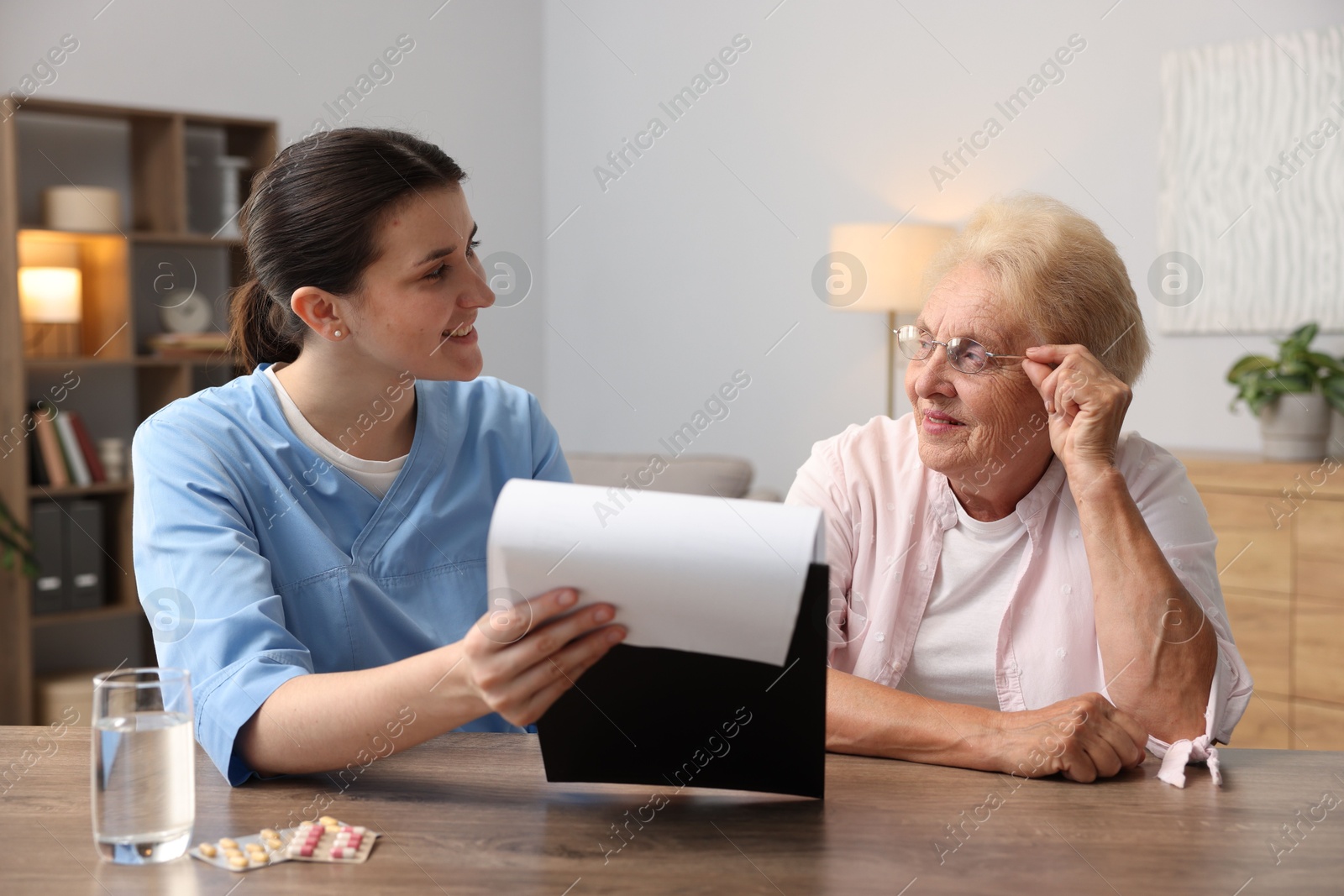 Nurse with clipboard examining senior woman at wooden table indoors. Home health care service Photo of Nurse with clipboard examining senior woman at wooden table indoors. Home health care service