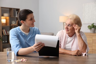 Photo of Nurse with clipboard examining senior woman at wooden table indoors. Home health care service
