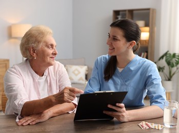 Photo of Nurse with clipboard examining senior woman at wooden table indoors. Home health care service
