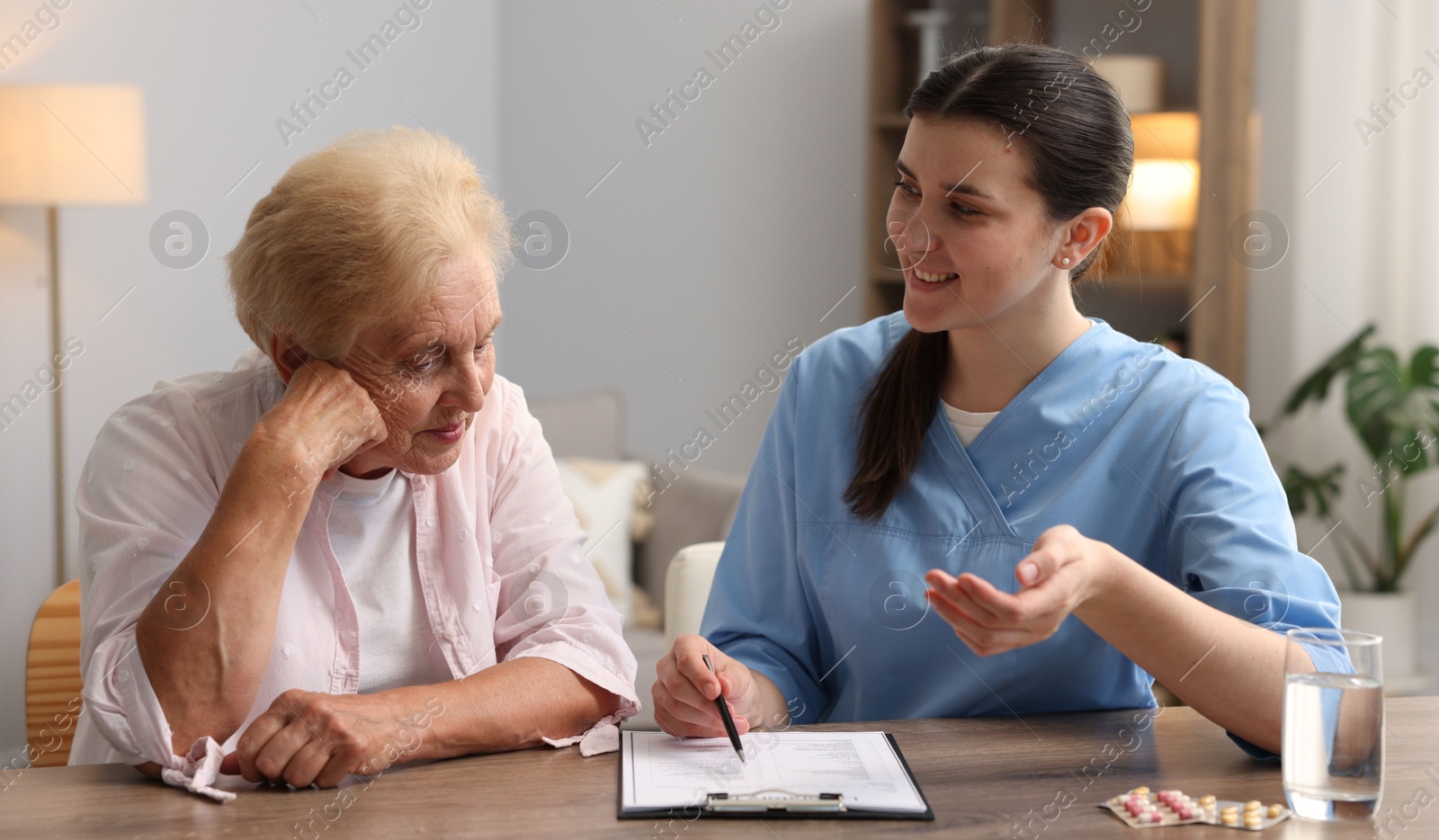Photo of Nurse with clipboard examining senior woman at wooden table indoors. Home health care service