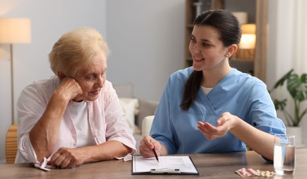 Photo of Nurse with clipboard examining senior woman at wooden table indoors. Home health care service