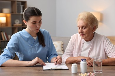 Photo of Nurse with clipboard examining senior woman at wooden table indoors. Home health care service
