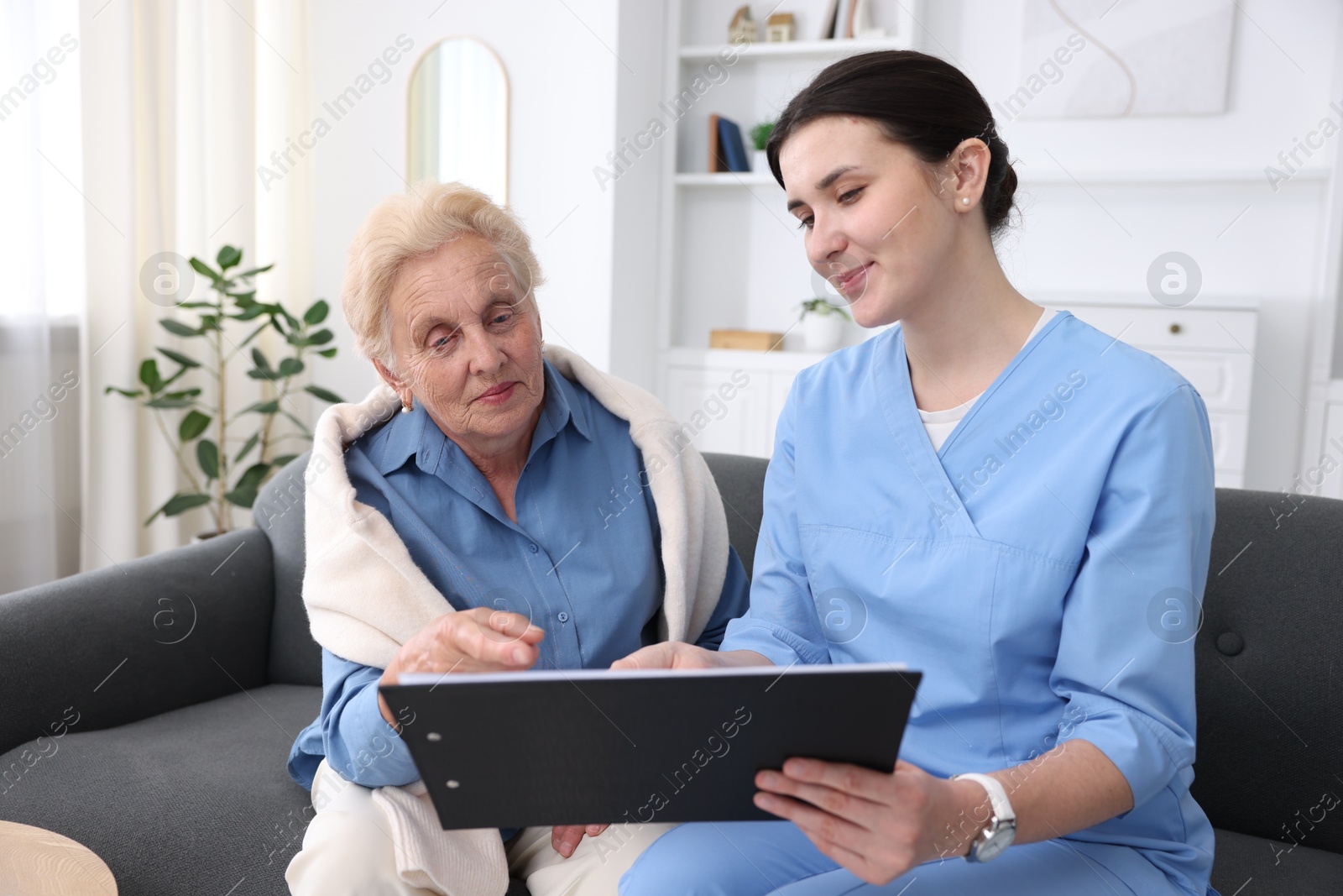 Photo of Nurse with clipboard examining senior woman indoors. Home health care service