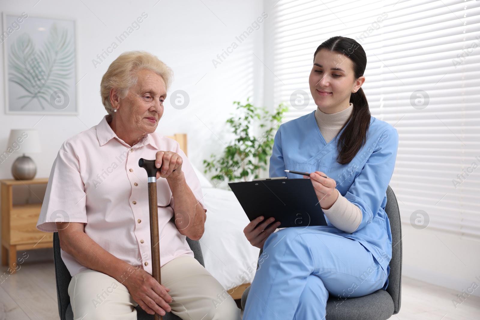 Photo of Nurse with clipboard examining senior woman indoors. Home health care service