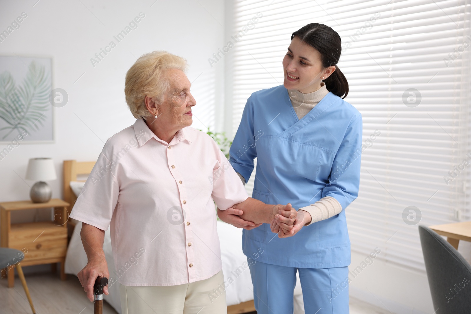 Photo of Nurse assisting senior woman with walking cane indoors. Home health care service