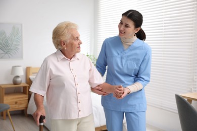 Photo of Nurse assisting senior woman with walking cane indoors. Home health care service