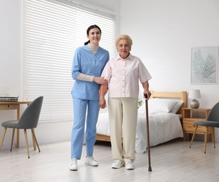 Photo of Nurse assisting senior woman with walking cane indoors. Home health care service