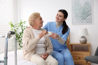 Photo of Nurse giving cup of drink to senior woman indoors. Home health care service