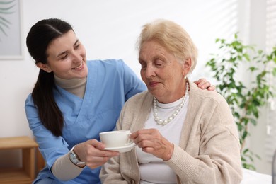 Photo of Nurse giving cup of drink to senior woman indoors. Home health care service