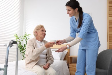 Photo of Nurse giving cup of drink to senior woman indoors. Home health care service