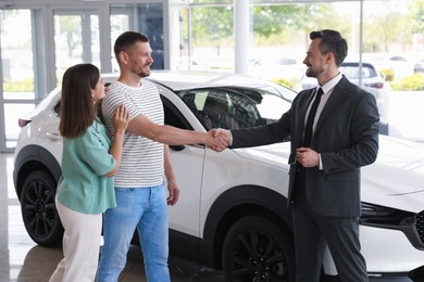 Photo of Salesman shaking hands with happy family near new car in salon