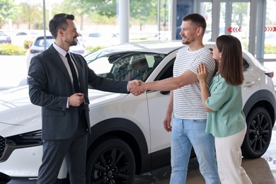 Photo of Salesman shaking hands with happy family near new car in salon