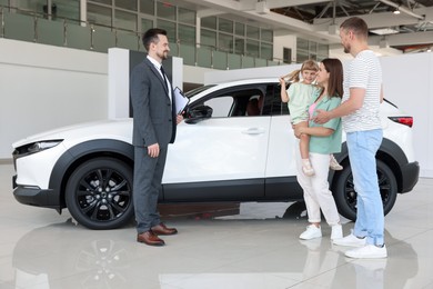 Photo of Smiling salesman with clipboard showing new car to family in salon