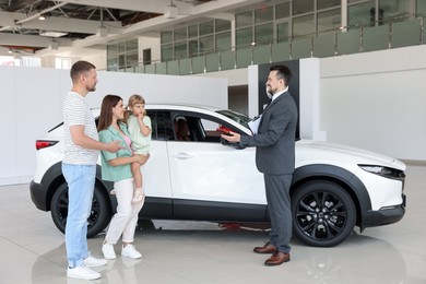 Photo of Smiling salesman with clipboard showing new car to family in salon