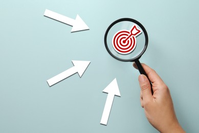 Image of Woman holding magnifying glass with target inside against light blue background, closeup. Arrows pointing at magnifier