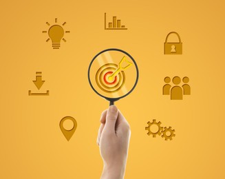 Image of Woman holding magnifying glass with target and arrow inside against orange background, closeup. Different icons around