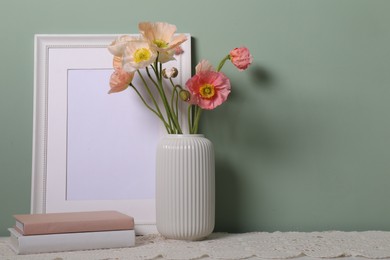 Photo of Beautiful poppy flowers in vase, books and empty frame on table near green wall, closeup