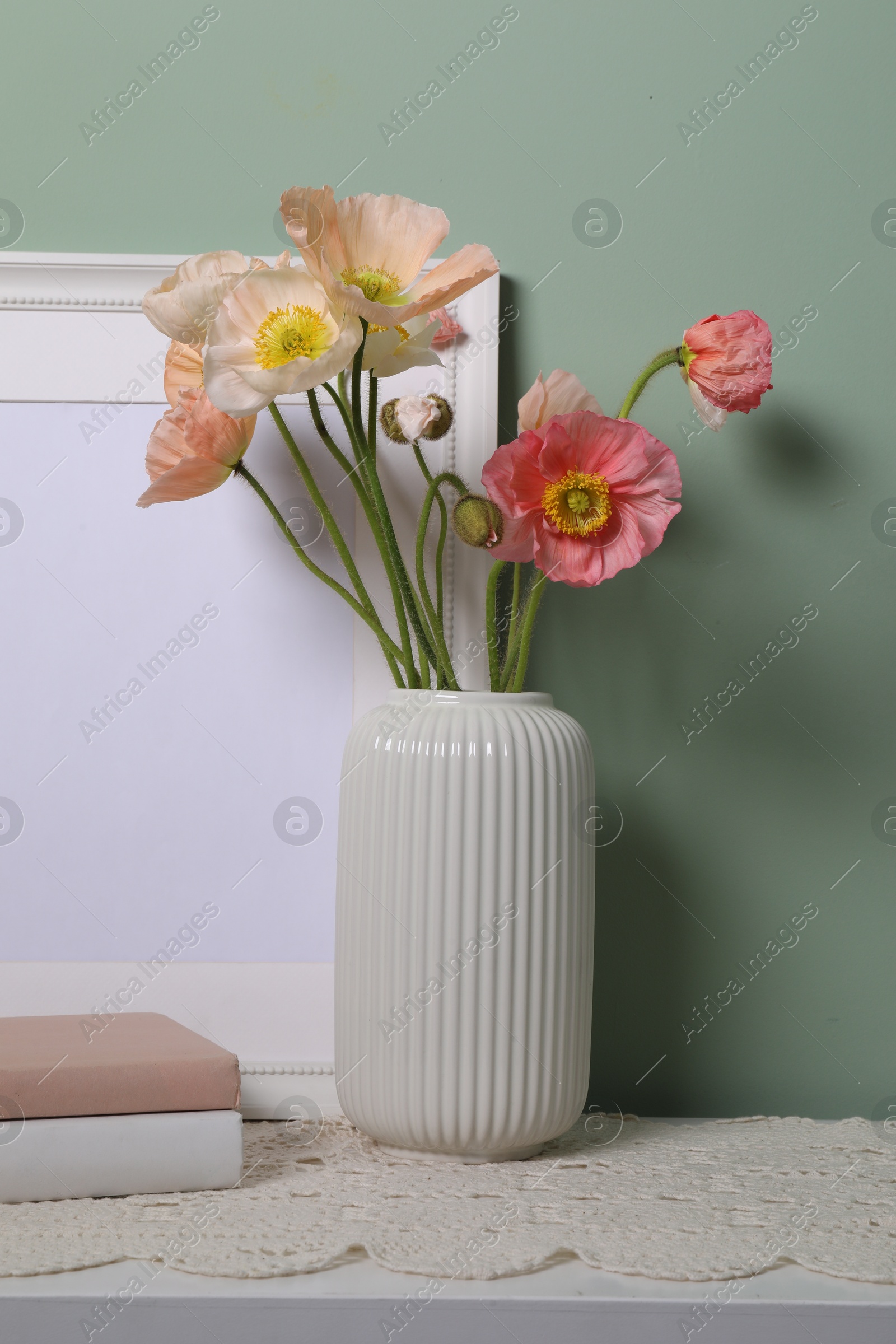Beautiful poppy flowers in vase, books and frame on light table near green wall, closeup Photo of Beautiful poppy flowers in vase, books and frame on light table near green wall, closeup