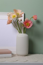 Photo of Beautiful poppy flowers in vase, books and frame on light table near green wall, closeup