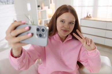 Photo of Beautiful teenage girl taking selfie by smartphone and showing peace sign on sofa at home