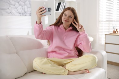 Photo of Beautiful teenage girl taking selfie by smartphone and showing peace sign on sofa at home