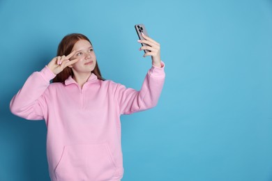 Photo of Beautiful teenage girl taking selfie by smartphone and showing peace sign on light blue background. Space for text