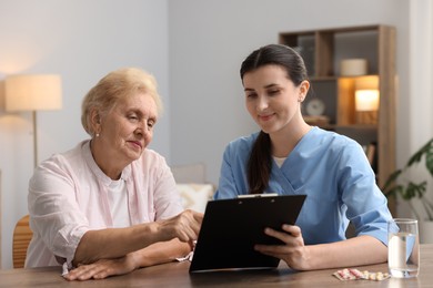 Photo of Nurse with clipboard examining senior woman at wooden table indoors. Home health care service