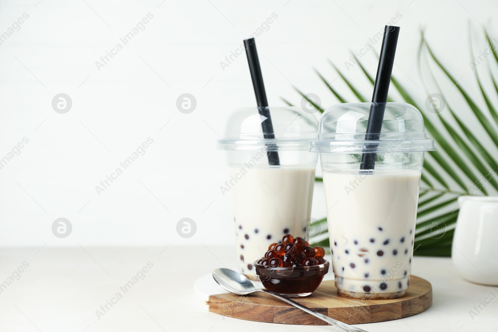 Photo of Tasty milk bubble tea in plastic cups, tapioca pearls and green leaves on beige table, space for text