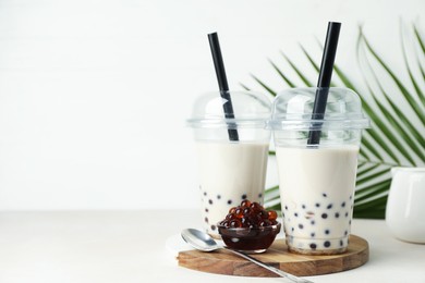 Photo of Tasty milk bubble tea in plastic cups, tapioca pearls and green leaves on beige table, space for text