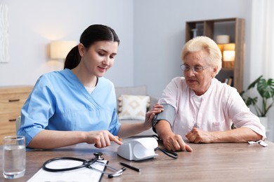 Photo of Nurse measuring patient's blood pressure at wooden table indoors. Home health care service