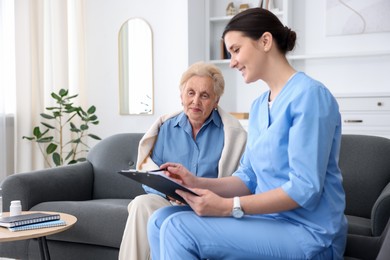 Photo of Nurse with clipboard examining senior woman indoors. Home health care service