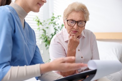 Photo of Nurse with clipboard examining senior woman indoors, closeup. Home health care service