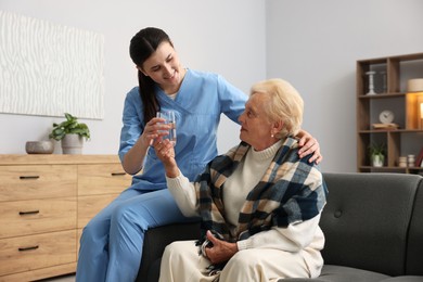 Photo of Nurse giving water to senior woman on sofa indoors. Home health care service