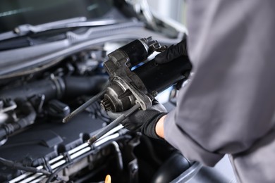 Photo of Auto part. Professional mechanic with starter motor near open hood of car indoors, closeup