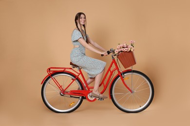 Photo of Smiling woman in dress riding bicycle with basket of flowers on beige background