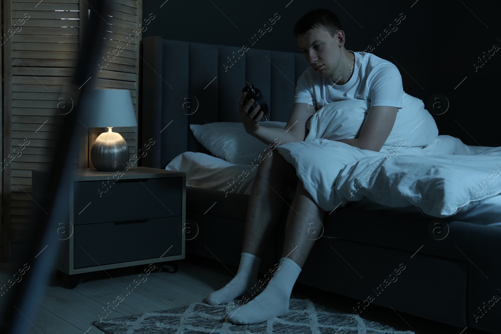 Frustrated man with alarm clock suffering from insomnia on bed indoors Photo of Frustrated man with alarm clock suffering from insomnia on bed indoors