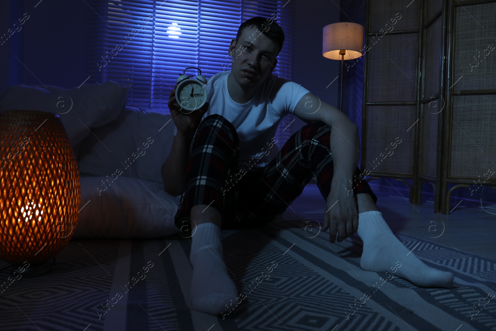 Frustrated man with alarm clock suffering from insomnia on floor indoors Photo of Frustrated man with alarm clock suffering from insomnia on floor indoors