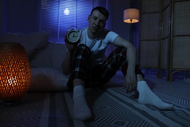 Photo of Frustrated man with alarm clock suffering from insomnia on floor indoors