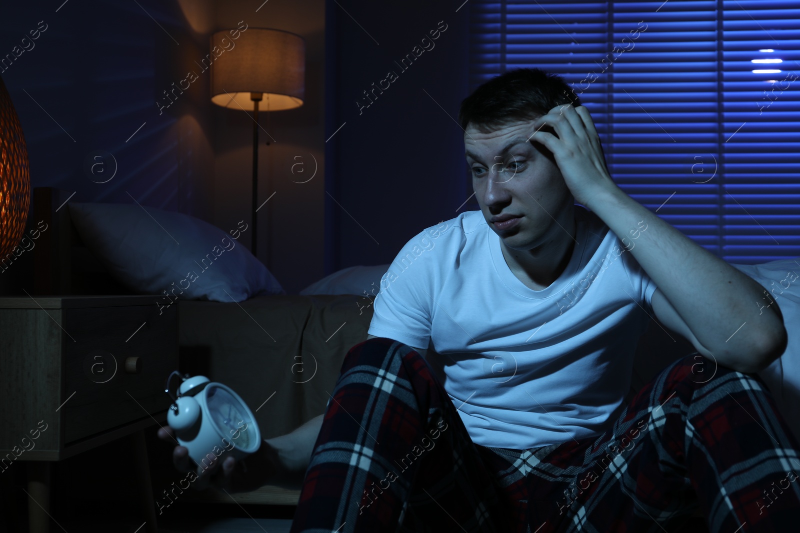 Frustrated man with alarm clock suffering from insomnia on floor indoors Photo of Frustrated man with alarm clock suffering from insomnia on floor indoors