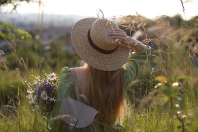 Photo of Woman with beautiful wildflowers, hat and backpack in meadow on sunny day, back view