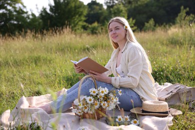 Photo of Woman with bouquet of wildflowers reading book on blanket in meadow