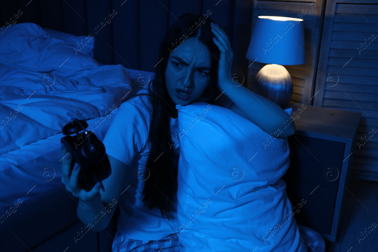 Photo of Frustrated woman with pillow and alarm clock suffering from insomnia on floor at home