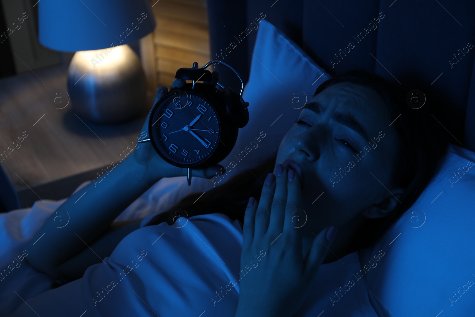 Frustrated woman with alarm clock suffering from insomnia in bed at home Photo of Frustrated woman with alarm clock suffering from insomnia in bed at home