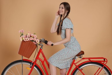 Photo of Smiling woman in dress riding bicycle with basket of flowers on beige background