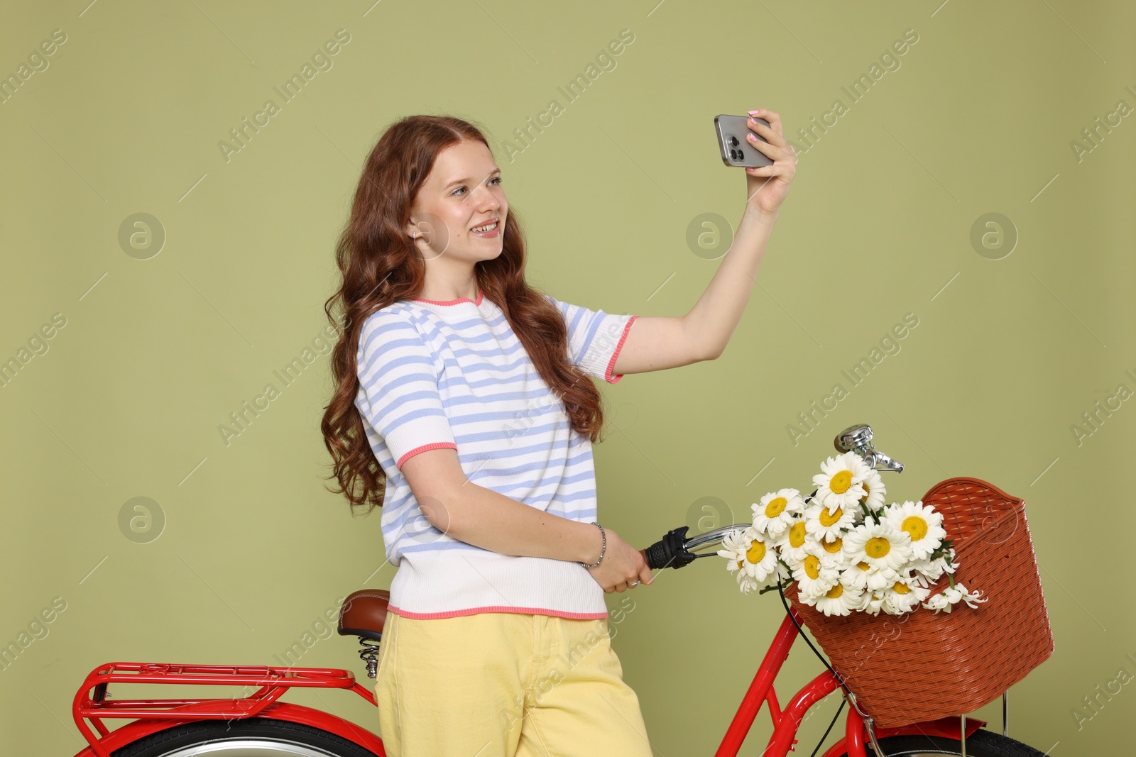 Smiling teenage girl taking selfie and bicycle with basket of flowers against green background Photo of Smiling teenage girl taking selfie and bicycle with basket of flowers against green background