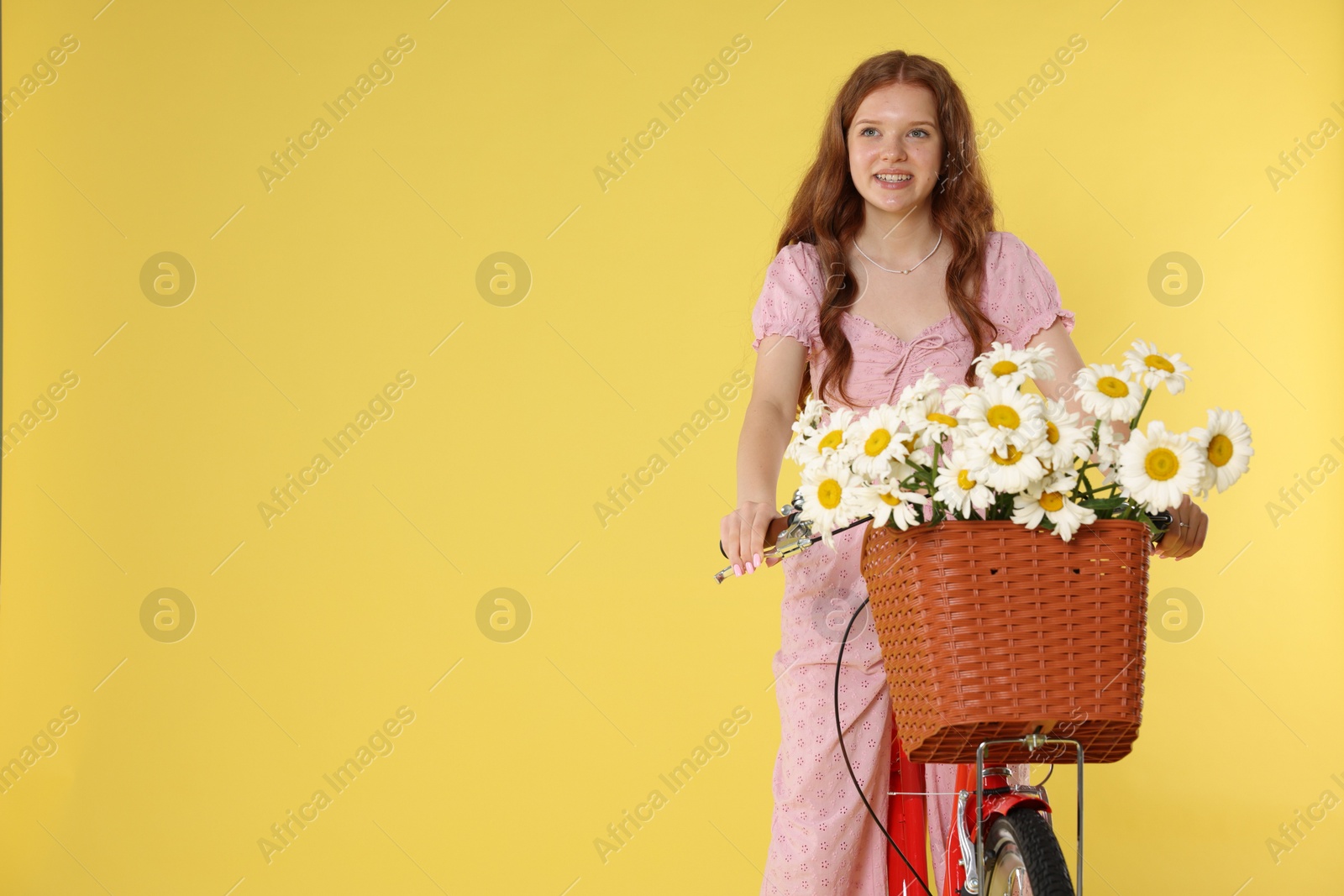 Smiling teenage girl riding bicycle with basket of flowers against yellow background. Space for text Photo of Smiling teenage girl riding bicycle with basket of flowers against yellow background. Space for text