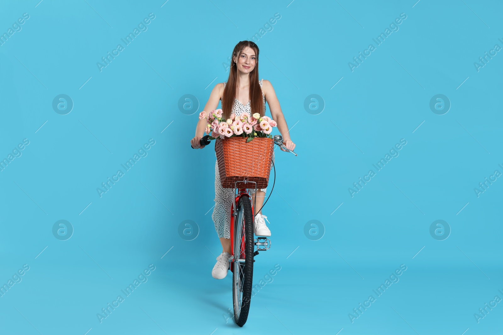 Beautiful woman riding bicycle with basket of flowers against light blue background Photo of Beautiful woman riding bicycle with basket of flowers against light blue background