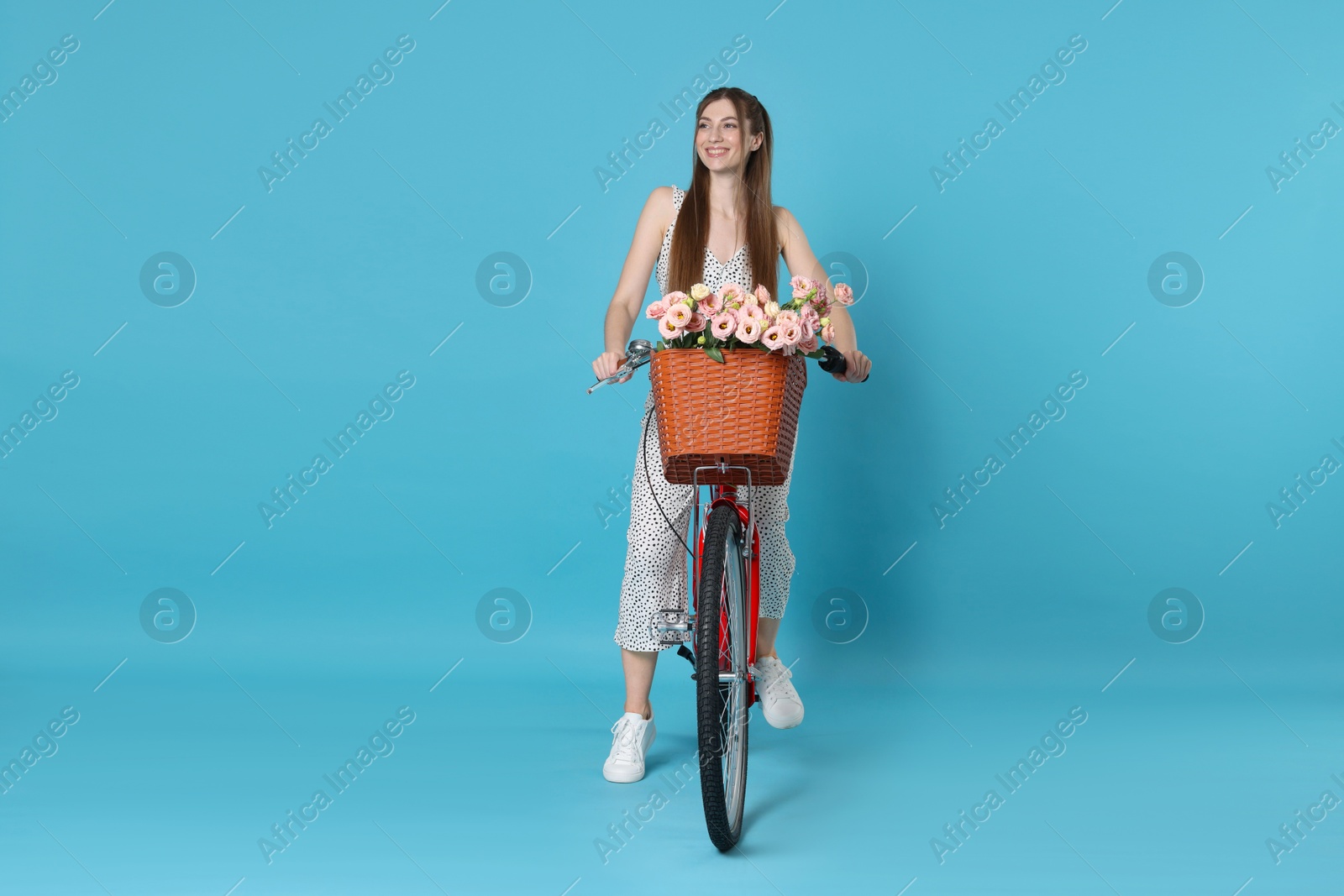 Smiling woman riding bicycle with basket of flowers against light blue background Photo of Smiling woman riding bicycle with basket of flowers against light blue background