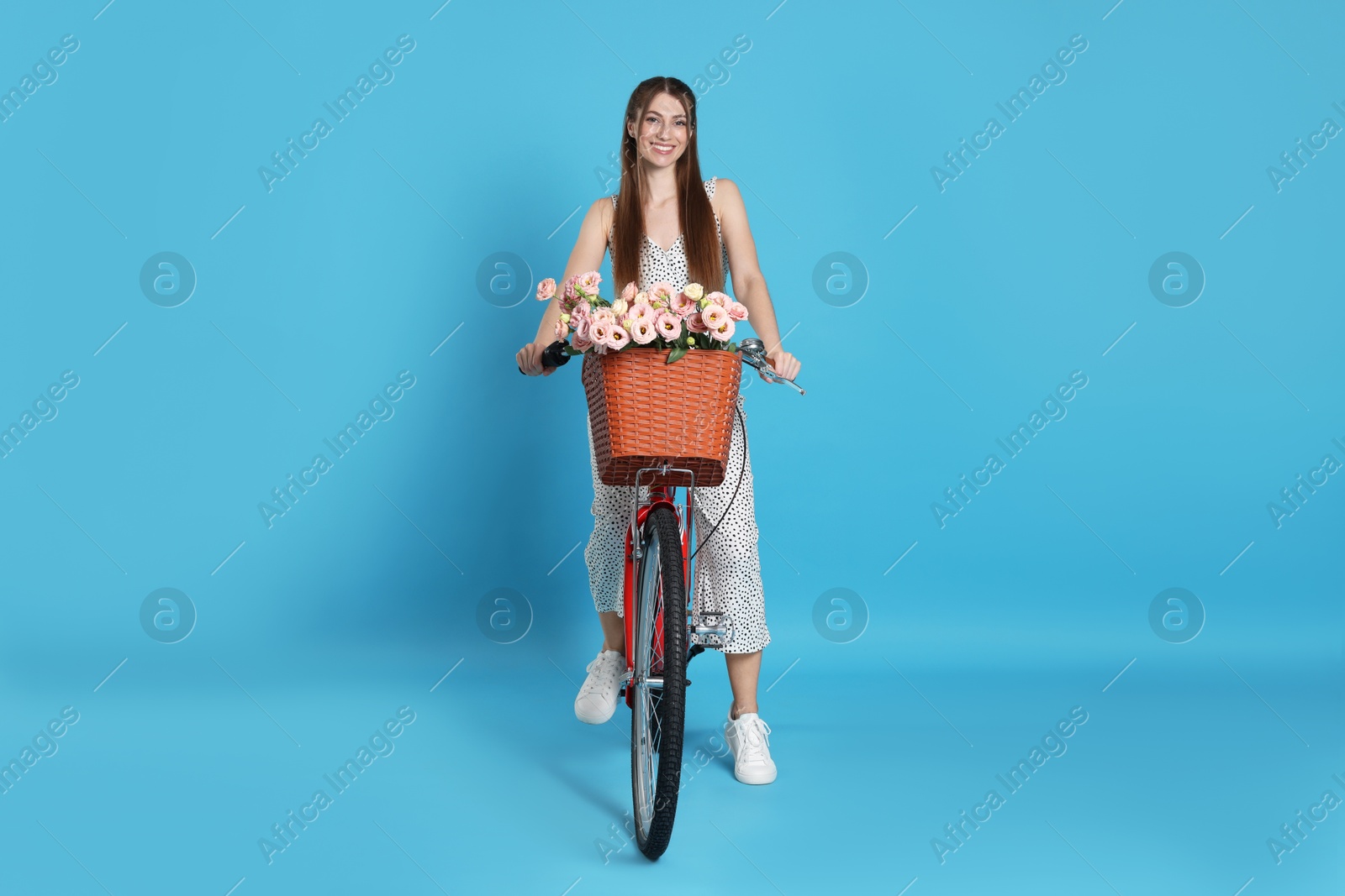 Smiling woman riding bicycle with basket of flowers against light blue background Photo of Smiling woman riding bicycle with basket of flowers against light blue background